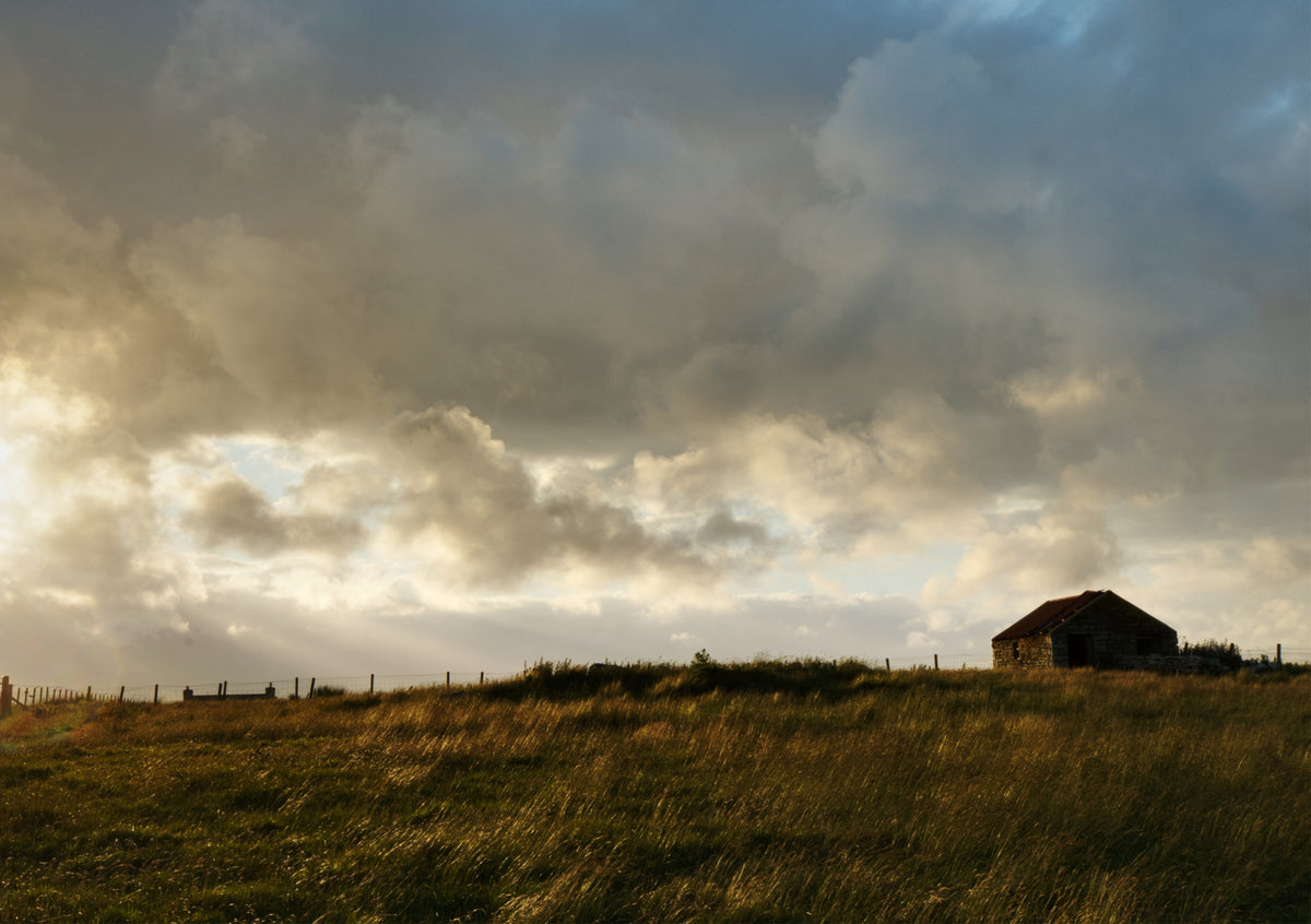 Fine Art Print eines Feldes der schottischen Highlands, mit dramatischen Wolken und rechts ein Haus mit Zaun